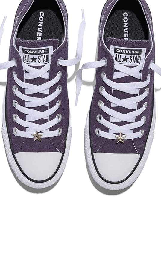 The image shows a pair of purple Converse All Star sneakers viewed from above, each with white laces and a star-shaped charm on the bottom lace. The shoes have the classic Converse logo on the tongue and black insoles with &ldquo;CONVERSE&rdquo; printed in white.