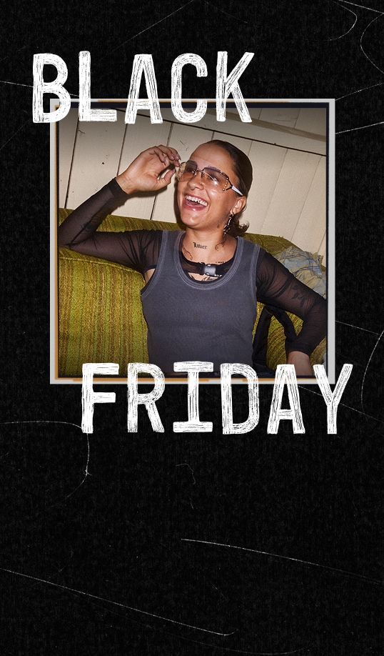 A person with slicked-back hair and clear glasses laughs while sitting on a green couch, wearing a dark sleeveless top over a mesh long-sleeve shirt. The photo is framed on a black background with large white distressed text reading &ldquo;BLACK FRIDAY.&rdquo;