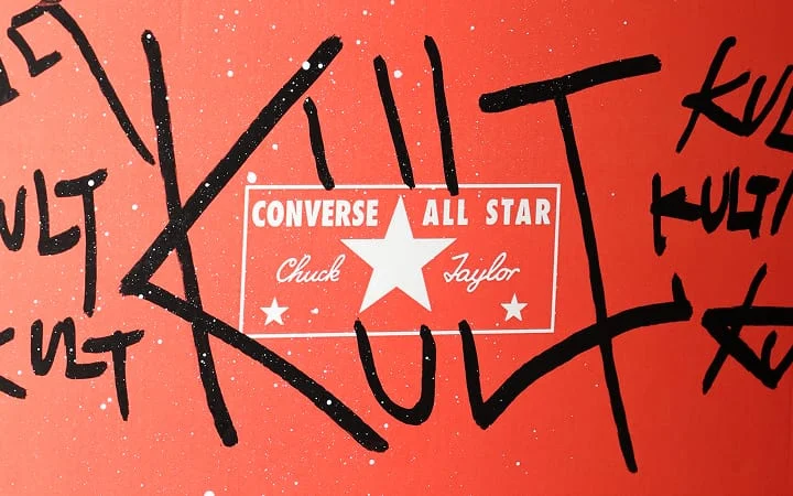 The image shows a red background with the white Converse All Star Chuck Taylor logo prominently displayed in the center. Surrounding the logo are large, black graffiti-style letters spelling out “KULT” multiple times.