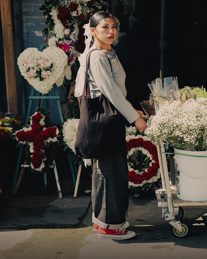 The image depicts a person standing outdoors next to a cart filled with white baby’s breath flowers, wearing a light gray ribbed long-sleeve top, loose dark pants rolled at the cuffs, red sneakers, and carrying a large black tote bag over one shoulder, with various floral wreaths including heart and cross shapes displayed in the background.