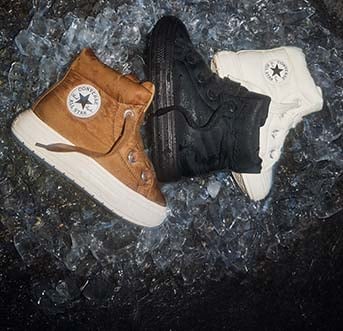 Three high-top Converse boots&mdash;brown, black, and white&mdash;are arranged together on a bed of crushed ice. Each boot shows the circular &ldquo;Converse All Star&rdquo; logo on the ankle panel.