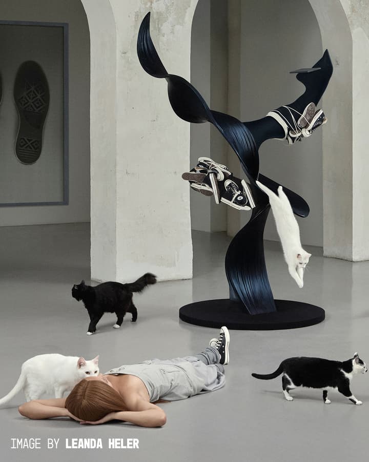 A woman lies on the floor in a minimalist gallery space surrounded by four cats and an abstract black sculpture adorned with sneakers, while one white cat leaps off the sculpture.