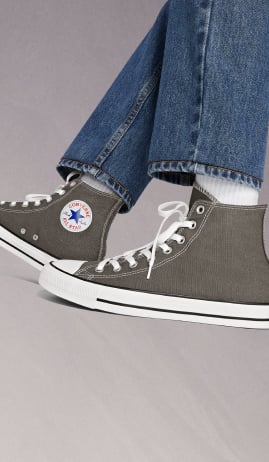 The image shows a person wearing gray Converse All Star high-top sneakers with white laces and white socks. They are also wearing blue denim jeans that slightly cover the tops of the shoes.