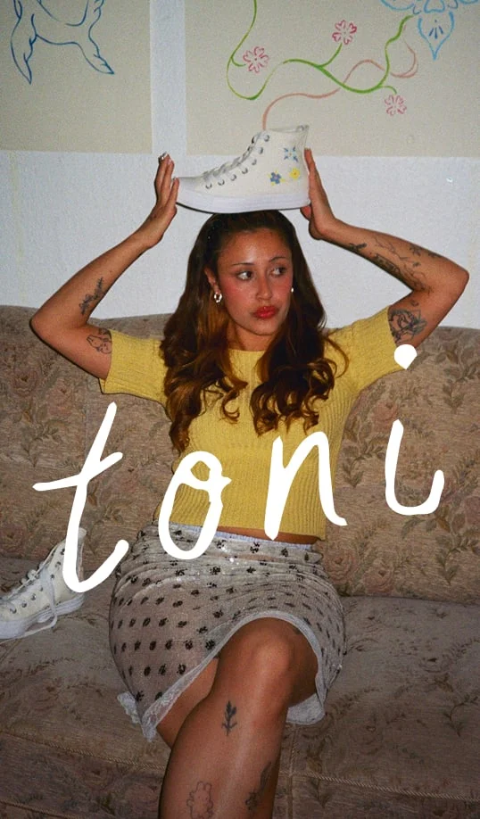 The image shows a person sitting on a patterned couch with their face blurred. They have long, wavy brown hair and are wearing a yellow short-sleeve top and a light-colored skirt with black polka dots or floral patterns. The person has multiple tattoos visible on both arms and legs. They are holding a white high-top sneaker decorated with small colorful flowers above their head using both hands. Another similar white sneaker is resting on the couch beside them. Behind the person, there are two pieces of wall art featuring simple line drawings of faces and floral designs in blue, green, pink, and red colors. The word &ldquo;toni&rdquo; is written prominently across the lower part of the image in large white handwritten-style text.