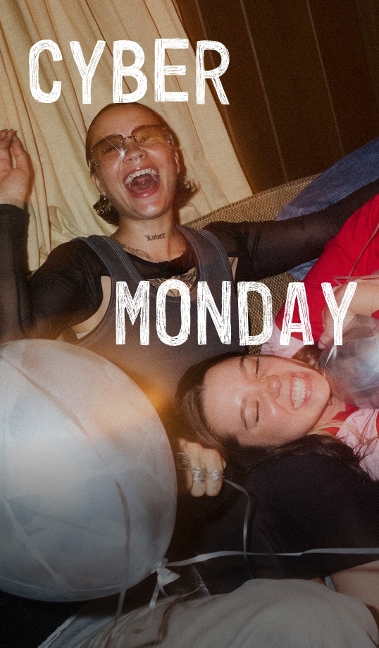 Two people laugh and lounge on a couch surrounded by balloons, one wearing sunglasses and a mesh top. White textured text over the photo reads “CYBER MONDAY.”