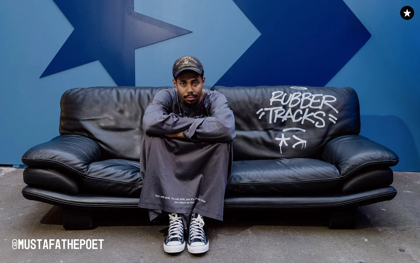 The image shows a person sitting cross-armed on a black leather couch with the words &ldquo;RUBBER TRACKS&rdquo; written in white graffiti-style text on the right side of the couch. The background features a blue wall with large star and arrow shapes, and the Instagram handle &ldquo;@MUSTAFATHEPOET&rdquo; is visible in the bottom left corner.