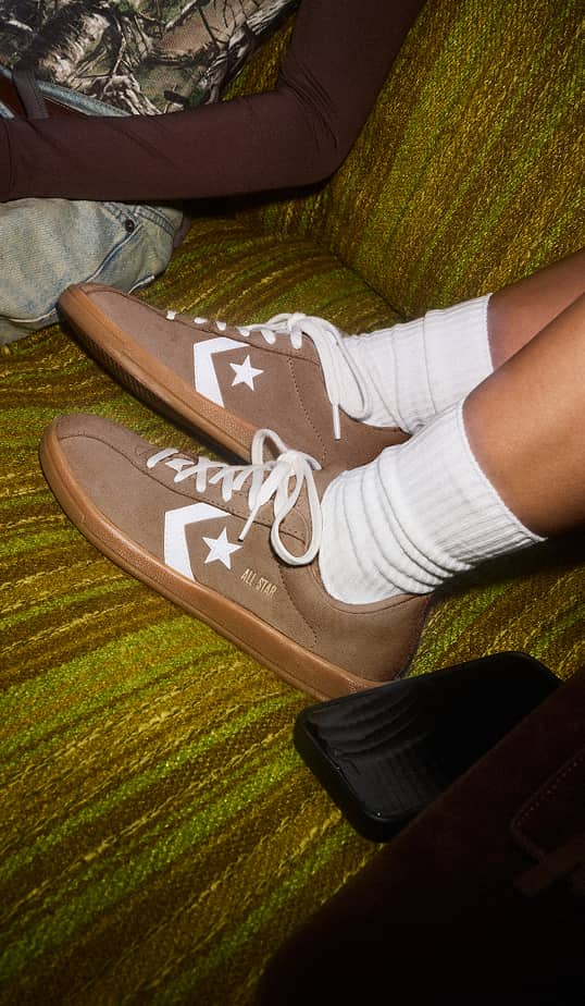 A pair of brown Converse-style sneakers with white star chevrons and white laces are worn with white crew socks, resting on a green textured couch. A person in jeans and a long-sleeve shirt sits nearby and a black phone lies beside the shoes.
