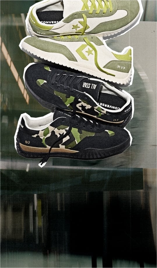 This image shows two pairs of Converse sneakers stacked vertically against a blurred background. The top pair is primarily white with olive green accents, including the laces, side stripe, and heel area. The word “MVP” is visible on the heel of this pair. The bottom pair features a black base with camouflage patterns in shades of green and beige on the sides. This pair has black laces and displays the text “ALL STAR” on the tongue and “DJ” near the heel. Both pairs have a sporty design with textured soles suitable for casual or athletic wear. The overall style suggests a modern, trendy look combining classic Converse branding with unique colorways and patterns.