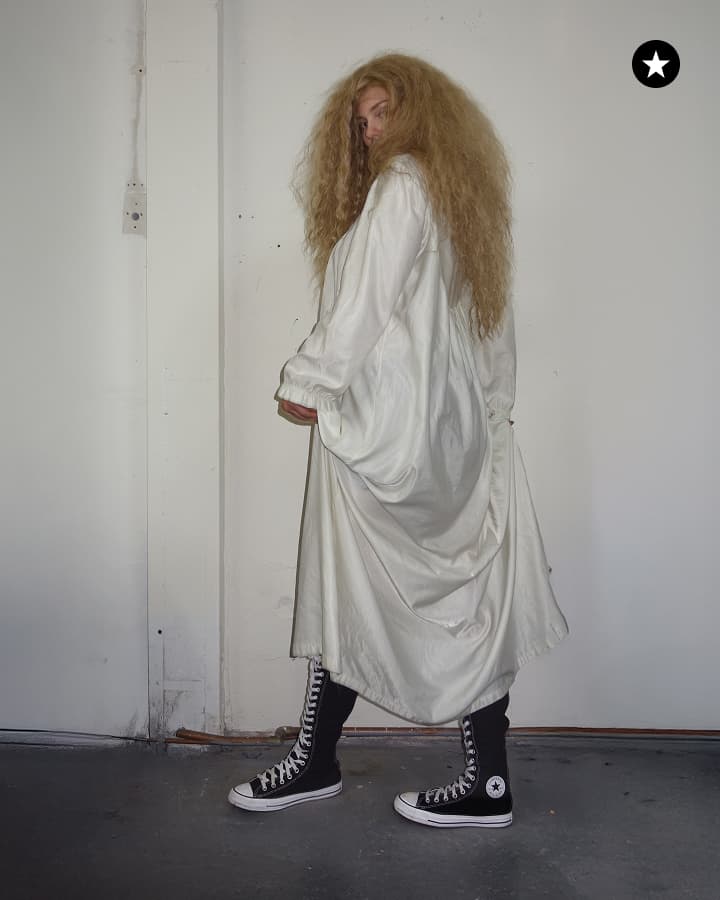A person with long, voluminous blonde hair is standing sideways against a plain white wall, wearing a loose white dress and tall black Converse-style high-top sneakers.
