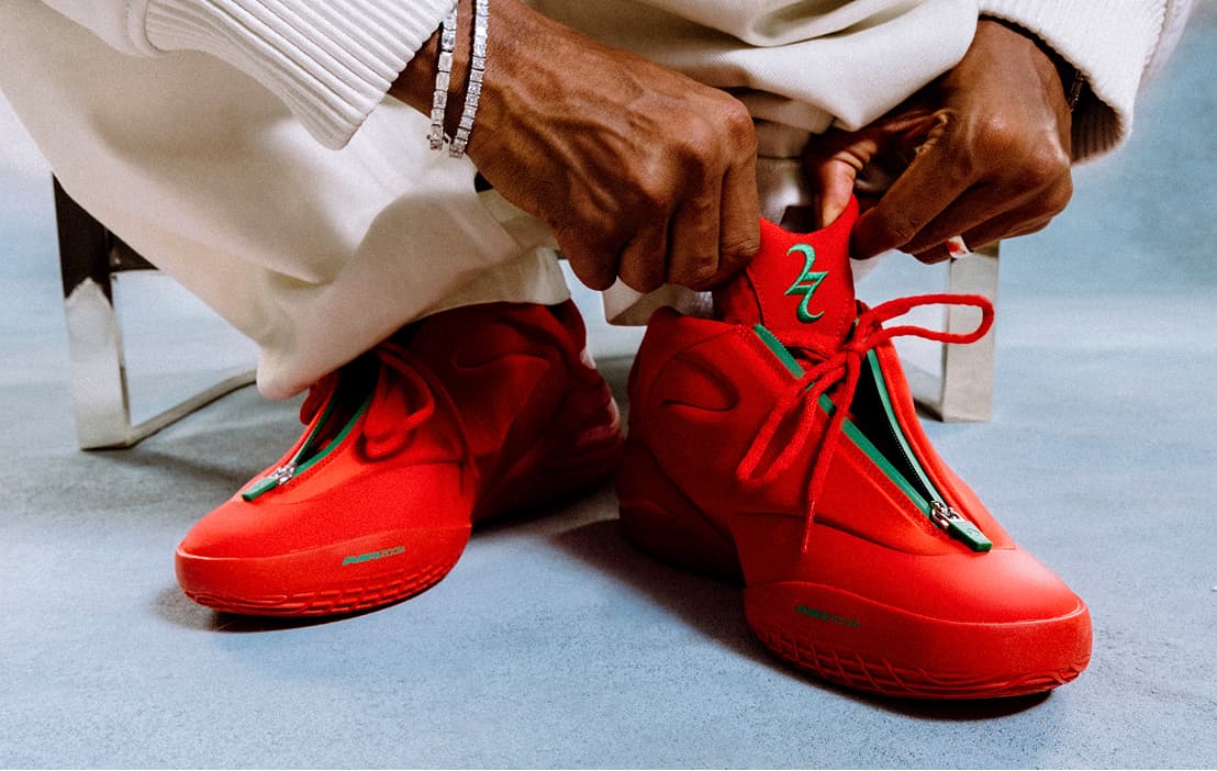 A person seated on a low chair is putting on bright red sneakers with green zipper accents and matching red laces. Their hands, wearing silver bracelets, adjust the tongue while loose white pants drape over the shoes on a light blue floor.