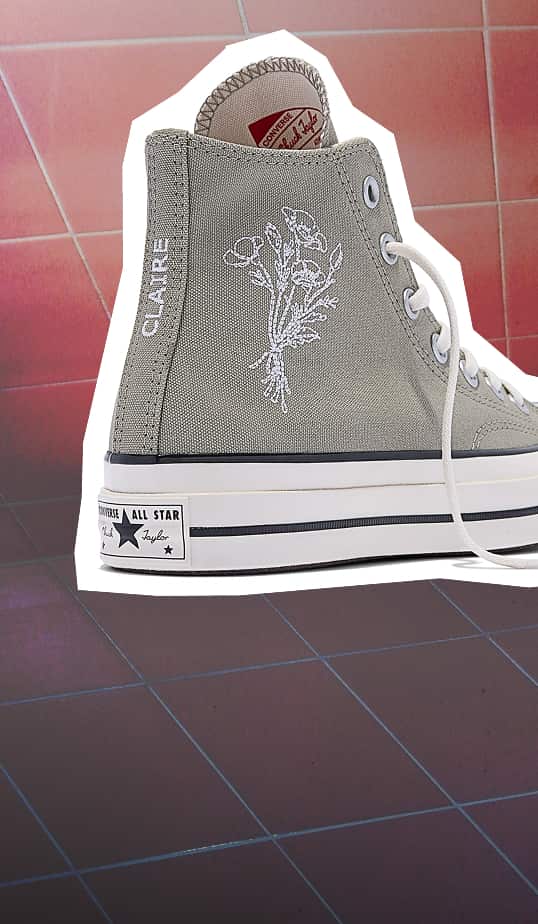 The image shows a close-up of a gray Converse Chuck Taylor All Star high-top sneaker with white laces, featuring an embroidered floral design and the name “CLAIRE” on the heel tab. The shoe is placed against a tiled floor background with a white cutout effect around it.