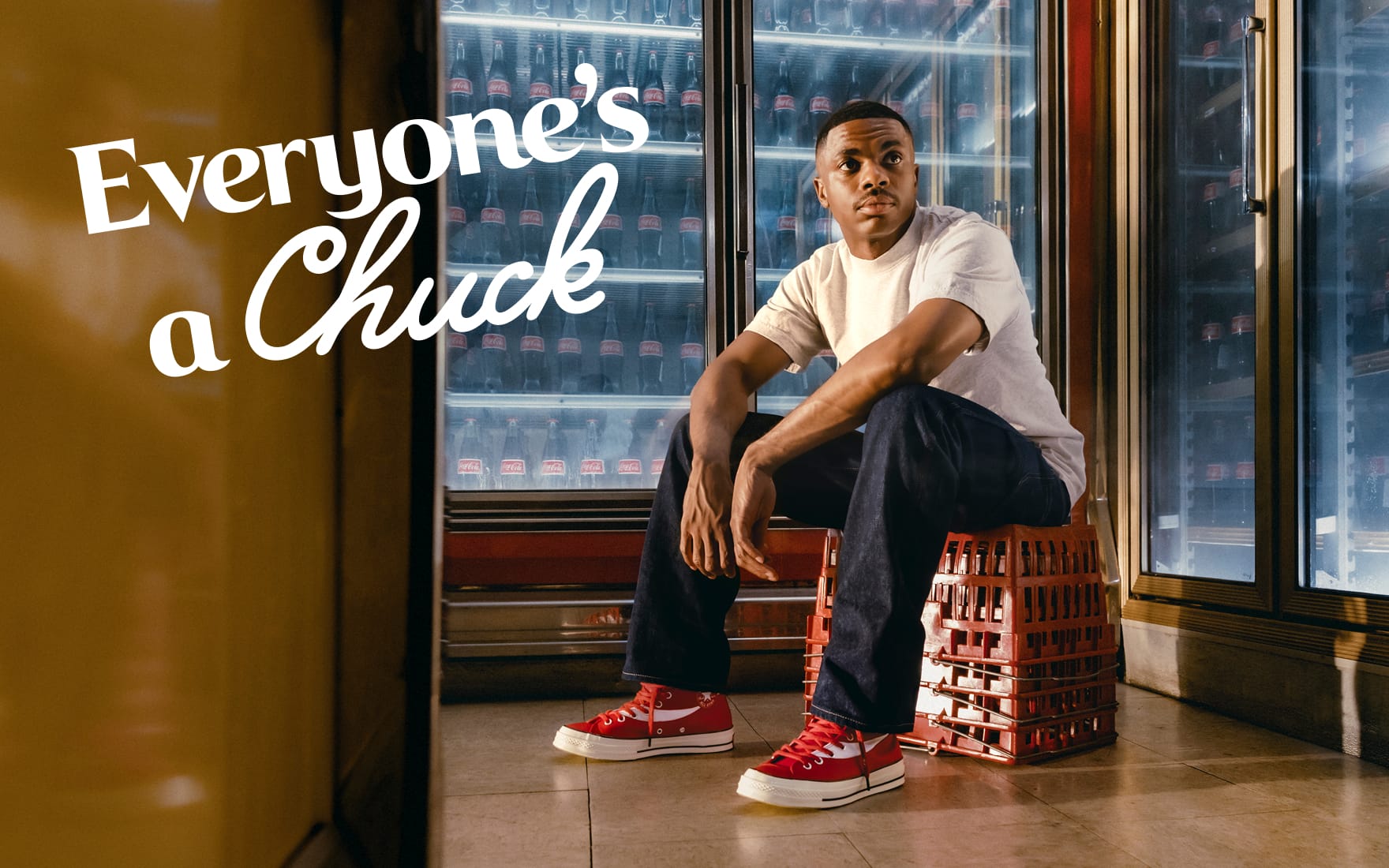 The image shows a person sitting on stacked red crates in front of refrigerated shelves filled with Coca-Cola bottles, wearing red Converse Chuck Taylor sneakers. The text &ldquo;Everyone&rsquo;s a Chuck&rdquo; is prominently displayed on the left side of the image.
