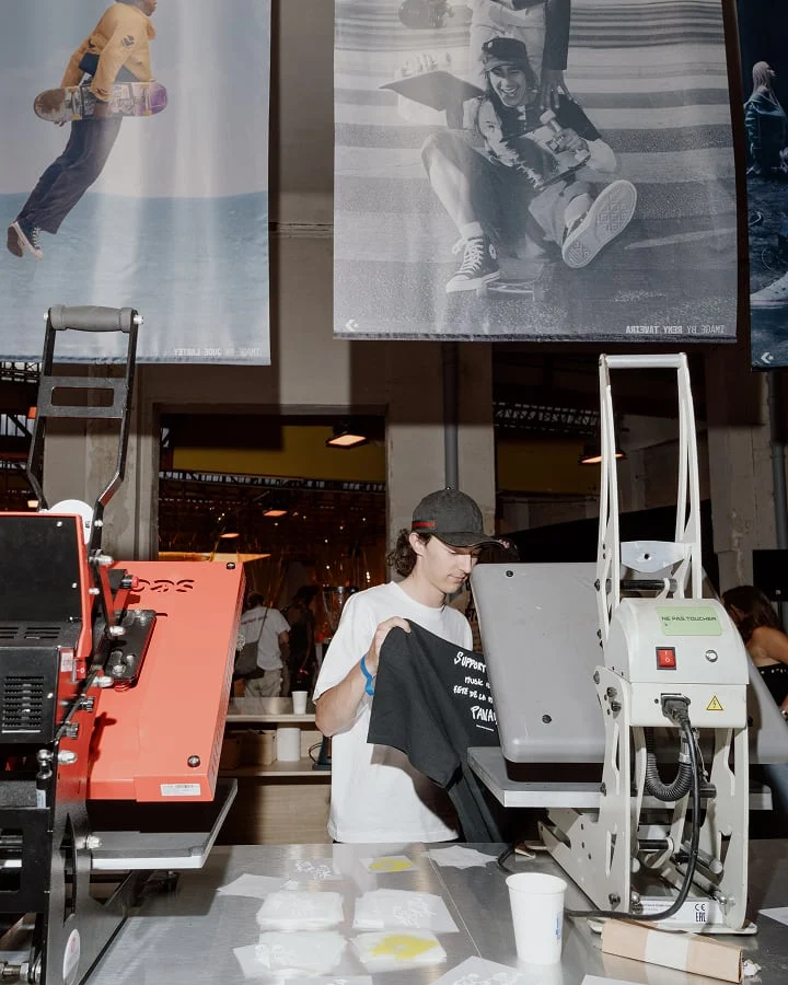 The image shows a person holding a black T-shirt in what appears to be a printing or merchandise booth with heat press machines on the table. Above them, there are large posters featuring skateboarding images hanging from the ceiling.
