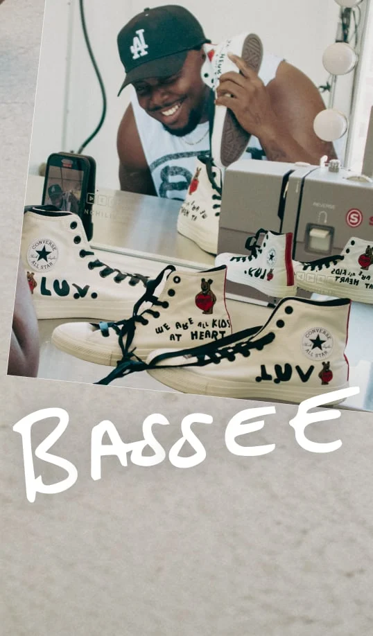 The image shows a person wearing a black cap with the letter &ldquo;A&rdquo; on it, holding a white Converse All Star high-top sneaker close to their face as if talking on it like a phone. The sneaker has custom artwork and text on it, including phrases like &ldquo;WE ARE ALL KIDS AT HEART&rdquo; and &ldquo;LUV.&rdquo; There is another similar sneaker placed in front of the mirror, reflecting both sneakers and part of the person&rsquo;s arm. A sewing machine is visible on the right side of the image near the mirror. The word &ldquo;BASSEE&rdquo; is handwritten in large white letters at the bottom of the photo. The overall setting appears to be indoors, possibly in a creative or personal workspace.