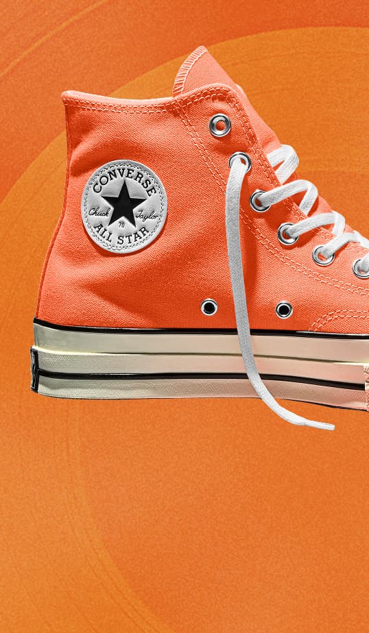 A bright orange Converse Chuck Taylor high-top sneaker is shown in close-up against a matching orange background. The shoe features white laces, metal eyelets, and the classic circular All Star logo on the ankle.