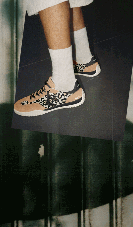 This image shows a close-up of a person&rsquo;s lower legs and feet wearing white crew socks and stylish sneakers. The sneakers have a distinctive design featuring leopard print patterns combined with beige and black colors. The person appears to be standing or slightly elevated against a dark, possibly tiled floor background. The overall style suggests a casual yet trendy look focused on the footwear.
