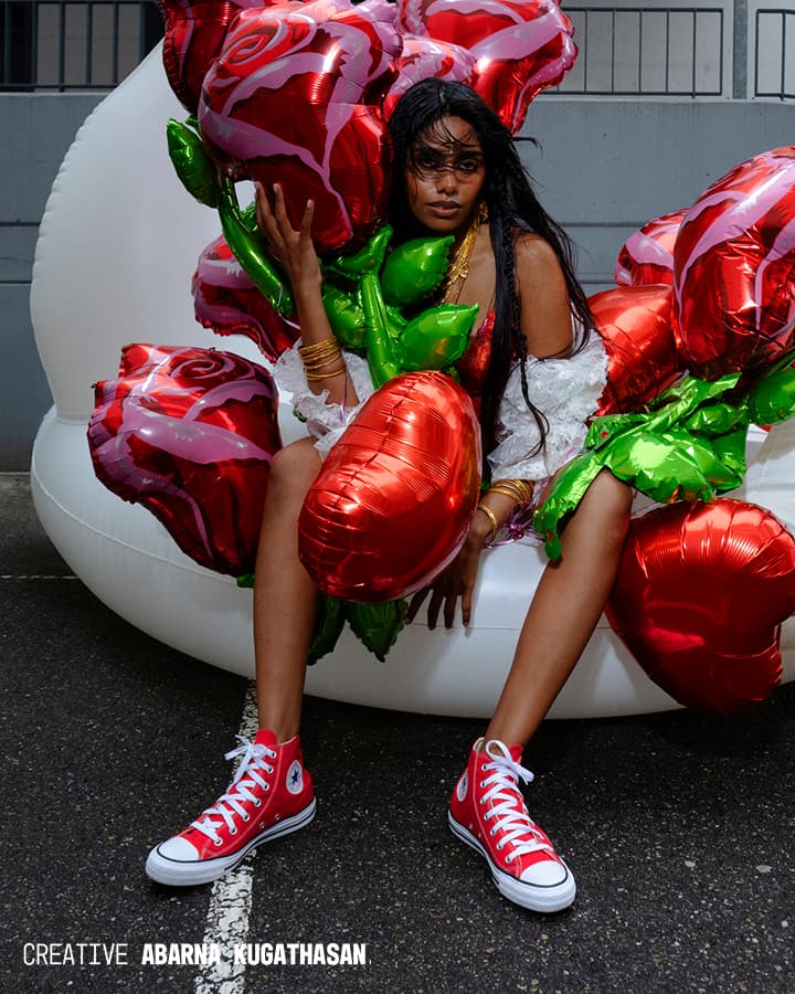 A person with long dark hair, wearing red high-top Converse sneakers and gold bracelets, sits on a large white inflatable chair outdoors while holding numerous shiny red heart-shaped balloons with green leaves.