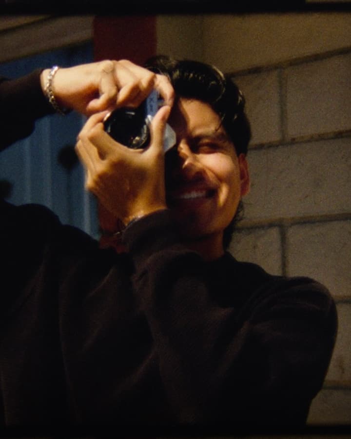 A person wearing a dark sweater and a silver bracelet is holding a camera up to their face, taking a photo against a background of tiled walls.