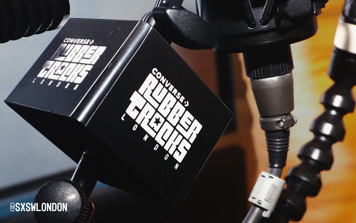 The image shows a close-up of a black microphone flag with the white text &ldquo;CONVERSE RUBBER TRACKS LONDON&rdquo; printed on it. In the bottom left corner, there is a social media handle &ldquo;@SXSWLONDON&rdquo; in white text.