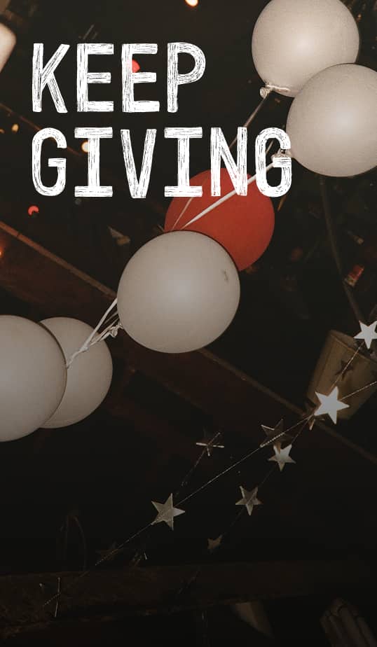 The image shows white and one red balloon cluster hanging against a dark ceiling with a string of small star decorations. Bold, white text at the top reads &ldquo;KEEP GIVING.&rdquo;