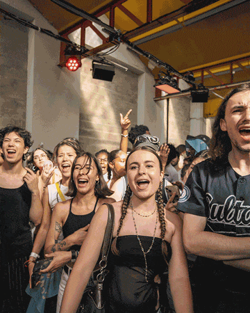 The image shows a group of people gathered indoors under a yellow ceiling with exposed beams and lighting equipment. Most individuals are casually dressed, some making hand gestures, suggesting a social or party atmosphere.