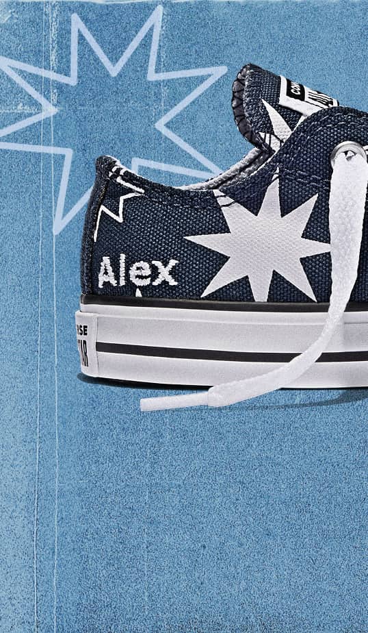The image shows a close-up side view of a dark blue sneaker with white starburst patterns on it. The shoe has white laces, and the name “Alex” is embroidered in white on the heel area. The sole is white with black stripes near the bottom edge.