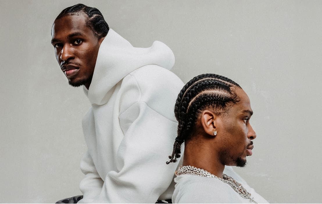 Two men sit back-to-back against a plain light background. One wears a white hooded garment and looks toward the camera, while the other with cornrows and chains faces sideways.