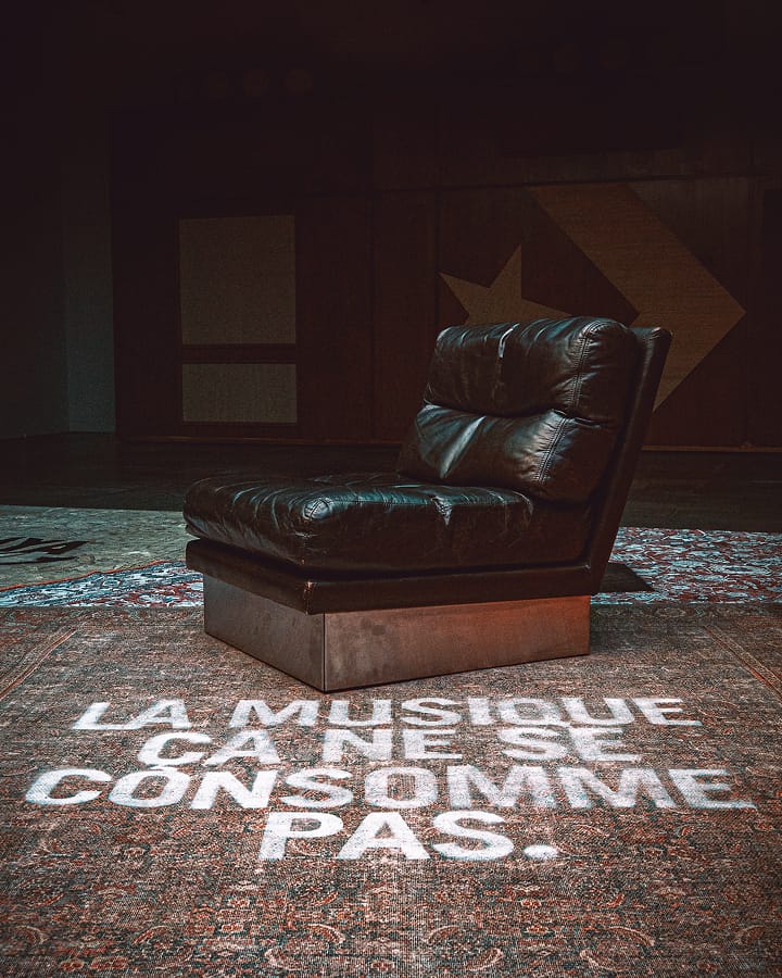 The image shows a dark leather chair placed on a patterned rug with the French text &ldquo;LA MUSIQUE &Ccedil;A NE SE CONSOMME PAS.&rdquo; projected or painted in white on the rug in front of it. The background is dimly lit, featuring wooden panels and geometric shapes.