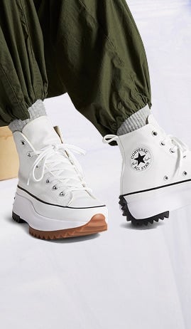 The image shows a pair of white high-top Converse All Star sneakers with chunky, rugged soles in different colors—brown on the front and black on the back. The shoes are worn with gray socks and olive green pants that are gathered at the ankles.