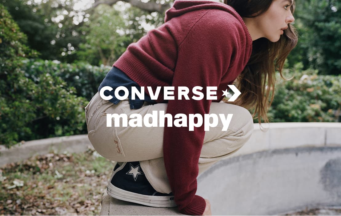 A person in a maroon hoodie and beige pants is crouched on a low concrete ledge outdoors, wearing black Converse sneakers with a star logo. Bold white text over the image reads “CONVERSE” with a star arrow symbol and “madhappy” beneath it.