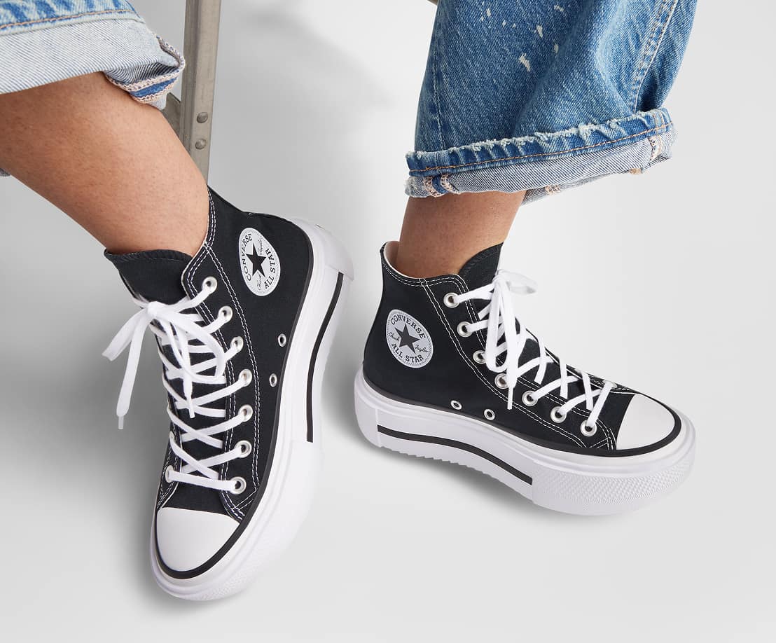 The image shows a person wearing black high-top Converse All Star sneakers with white laces and thick white soles. The person is also wearing rolled-up blue jeans, and their feet are slightly lifted off the ground against a plain light background.