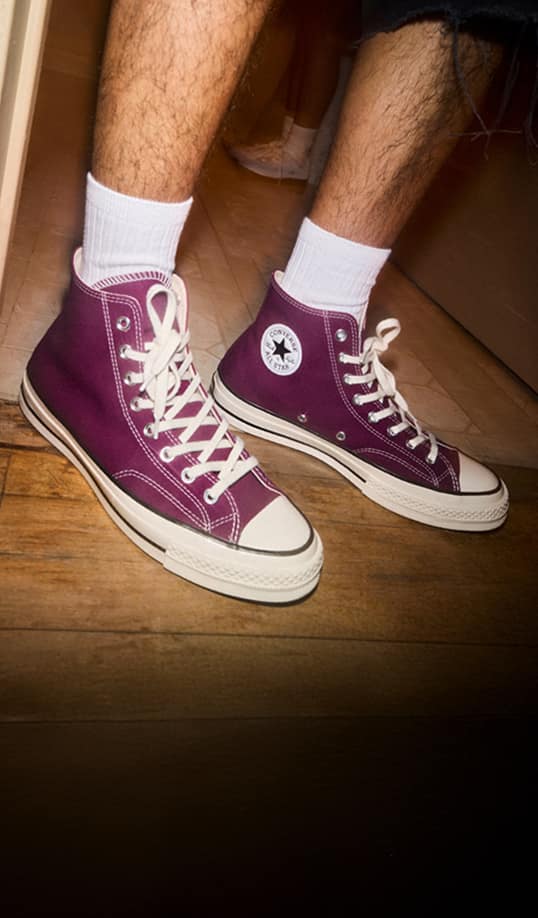 A pair of purple high-top Converse sneakers with white laces and soles are shown on a wooden floor, worn with white crew socks. The photo focuses on the lower legs and feet, with warm indoor lighting and a slight shadow cast behind them.