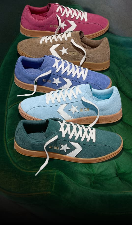 Five low-top Converse &ldquo;All Star&rdquo; sneakers in pink, brown, blue, light blue, and teal are neatly stacked on a green velvet surface. Each shoe features white laces, a white star-chevron logo, and a gum-colored sole.