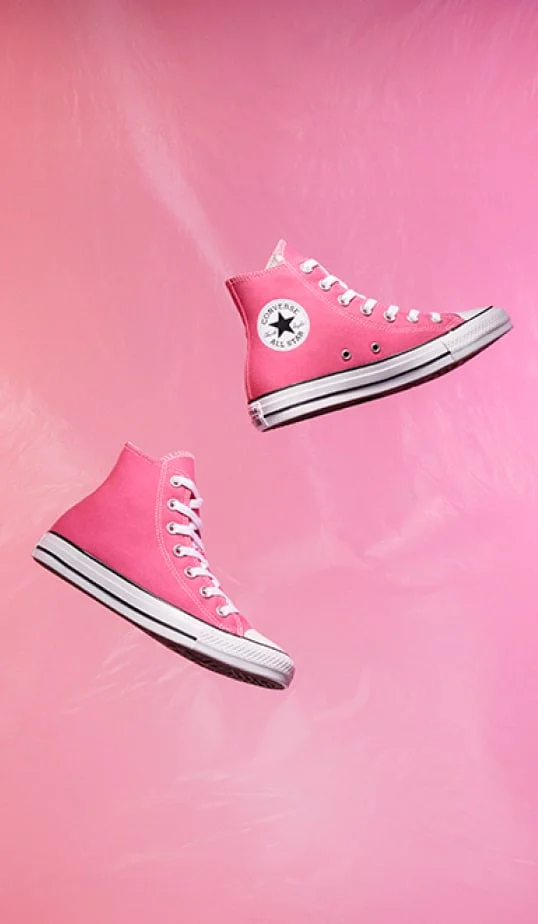 The image shows two pink high-top Converse sneakers floating against a matching pink background. One shoe displays the classic Converse All Star logo, and both have white laces and soles with black stripes.