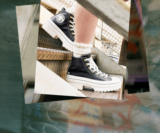 The image shows a close-up of a person&rsquo;s legs and feet wearing black high-top sneakers with white soles and laces. The shoes have a distinctive circular logo on the side, resembling the Converse All Star design. The person is standing on metal stairs with a textured surface, possibly outdoors or in an industrial setting. They are also wearing white socks that are slightly scrunched down above the shoes. The background includes metal railings and some structural elements, giving an urban or utilitarian feel to the scene.