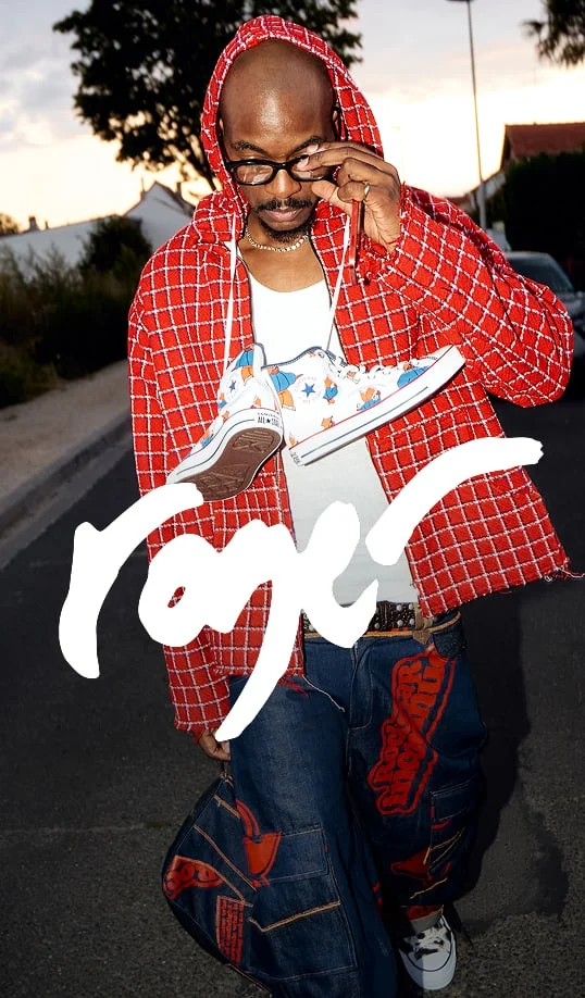 The image shows a person standing outdoors on a street during what appears to be either early morning or late afternoon, given the lighting. The individual is wearing a red hooded jacket with a white grid pattern over a white shirt. They are holding up a pair of colorful sneakers featuring cartoon-like designs and stars by their laces in front of their chest. The person is also dressed in loose-fitting blue jeans with large red graphic prints on them and white sneakers on their feet. There is bold, stylized white text across the lower part of the image that reads &ldquo;roger.&rdquo; The background includes some trees, houses, and parked cars along the road. The person&rsquo;s face is blurred for privacy.