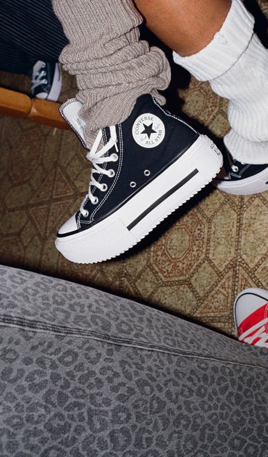  The image shows a close-up of a foot wearing a black Chuck Taylor All Star Double Stack Platform sneaker with thick white soles and white laces. The person is also wearing beige leg warmers scrunched down over the shoe and white socks underneath, with part of another red sneaker visible in the background.