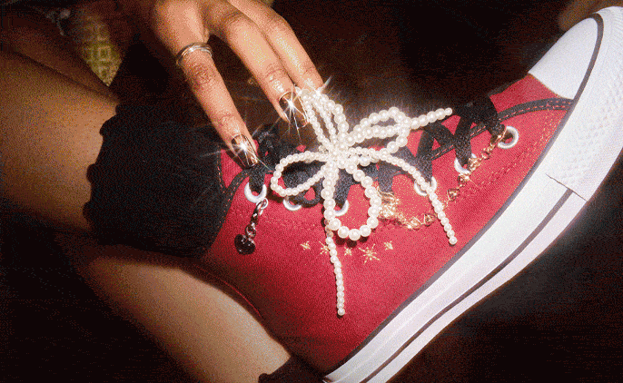 A close-up of a red high-top sneaker embellished with a pearl bow lace, gold star embroidery, and a small charm. A hand with long manicured nails and rings is adjusting the laces.