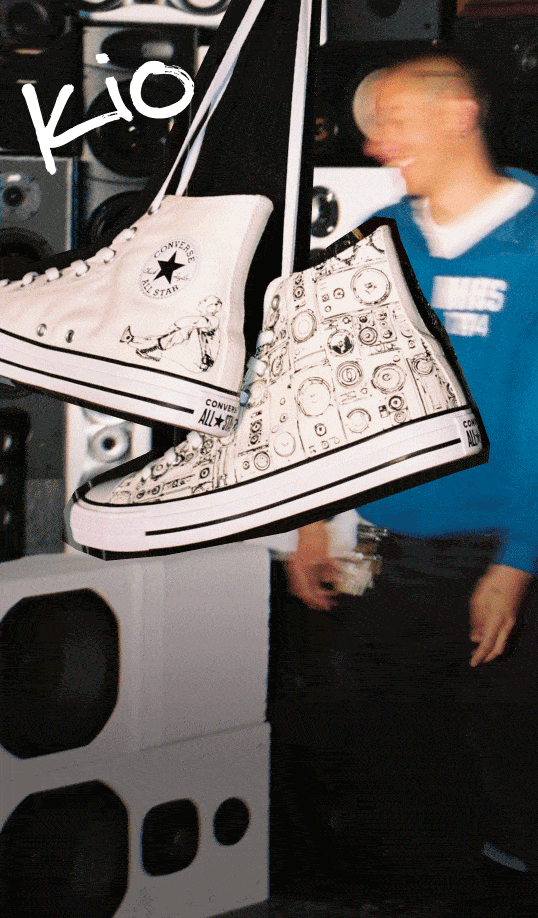 The image shows a pair of white Converse All Star high-top sneakers hanging by their laces in the foreground. One shoe features a simple design with the classic Converse logo and an illustration of a person sitting, while the other shoe is decorated with various black-and-white sketches resembling speakers or audio equipment. 