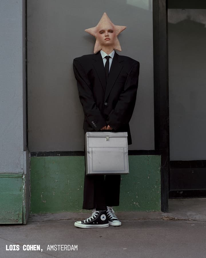 A person with a star-shaped object obscuring their face is standing against a two-tone gray and green wall, dressed in an oversized black suit, white shirt, black tie, holding a large light gray briefcase, and wearing black Converse high-top sneakers.