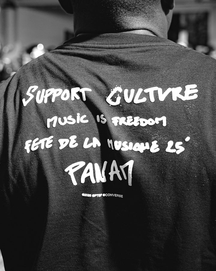 The image shows the back of a person wearing a black t-shirt with white handwritten-style text promoting culture and music. The text reads &ldquo;SUPPORT CULTURE,&rdquo; &ldquo;MUSIC IS FREEDOM,&rdquo; &ldquo;FETE DE LA MUSIQUE 25&rsquo;,&rdquo; and &ldquo;PANAM,&rdquo; along with a small Converse logo at the bottom.