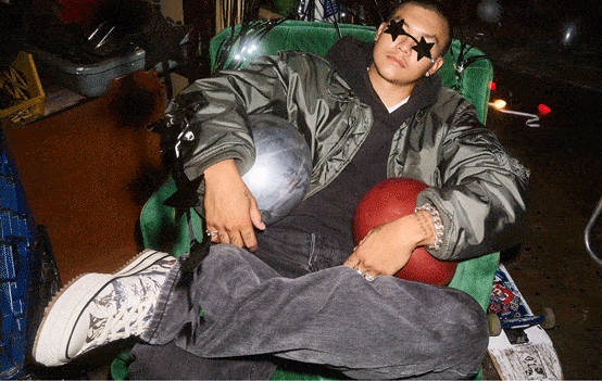 A person wearing a shiny bomber jacket and star-shaped sunglasses is reclining in a green armchair with legs crossed. They hold a silver and a red ball, and wear patterned high-top sneakers and multiple rings.