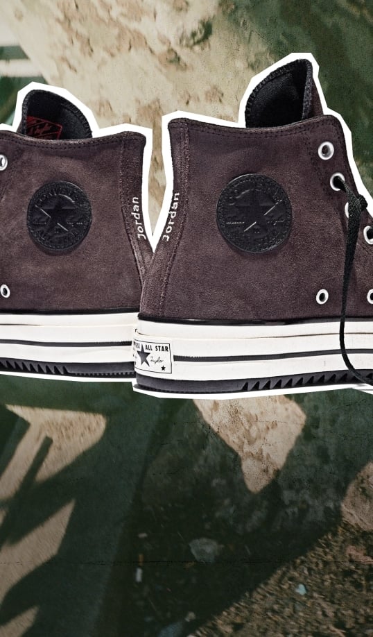 This image shows a close-up view of the back and side portions of a pair of high-top Converse sneakers. The shoes are dark brown or black suede with white rubber soles featuring black stripes. The iconic circular Converse All Star logo patch is visible on the ankle area of each shoe, but it appears to be in a dark tone matching the shoe color, making it less prominent. On the heel tab area between the two shoes, there is white text that reads &ldquo;jordan&rdquo; and &ldquo;lapoet,&rdquo; suggesting some form of collaboration or customization. The laces are black, and one lace is loosely hanging on the right shoe. The background appears to be an outdoor surface with shadows cast across it, possibly concrete or pavement with some dirt or debris scattered around.