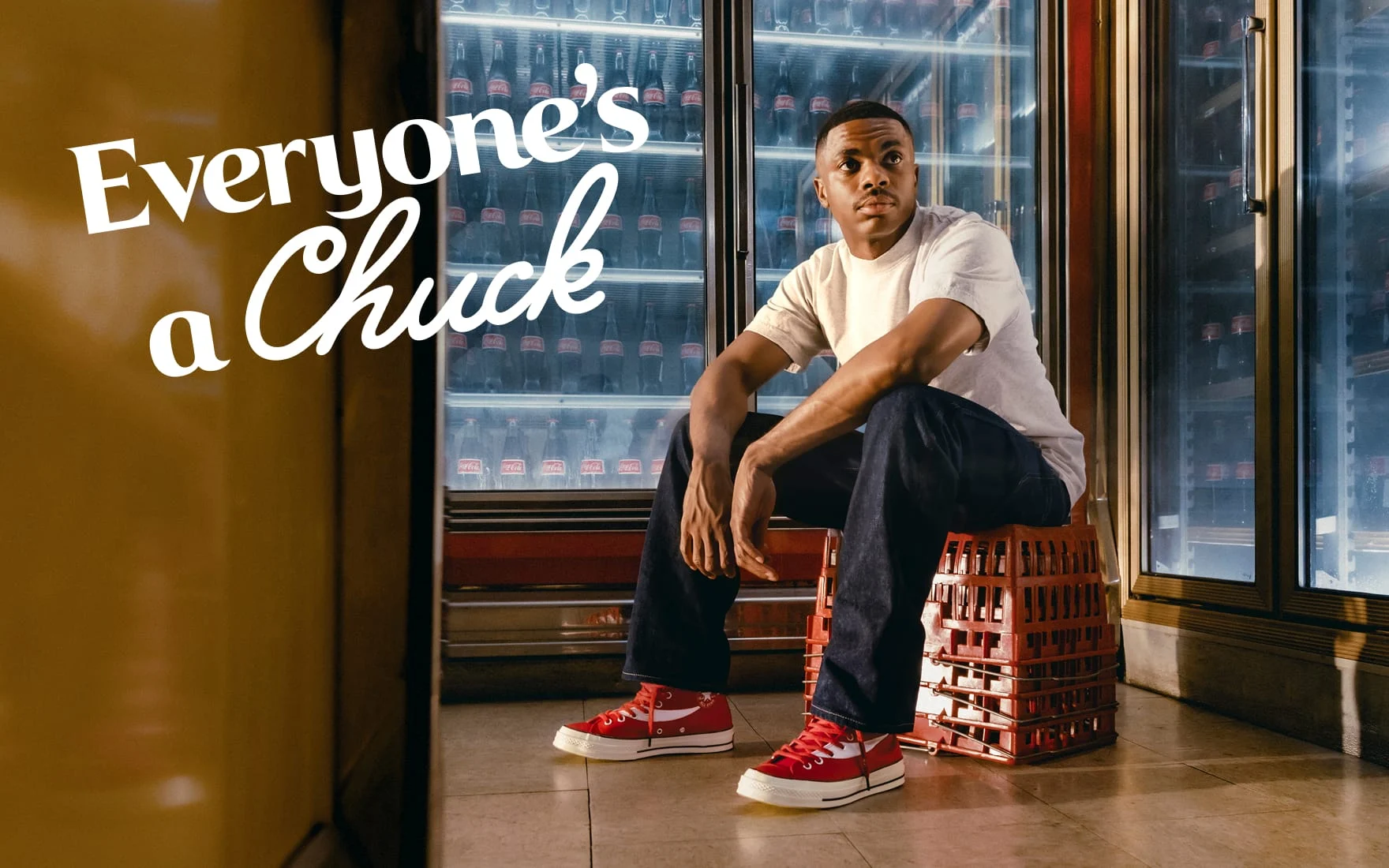 The image shows a person sitting on stacked red crates in front of refrigerated shelves filled with Coca-Cola bottles, wearing red Converse Chuck Taylor sneakers. The text &ldquo;Everyone&rsquo;s a Chuck&rdquo; is prominently displayed on the left side of the image.