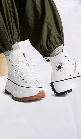 The image shows a pair of white high-top Converse All Star sneakers with chunky, rugged soles in different colors&mdash;brown on the front and black on the back. The shoes are worn with gray socks and olive green pants that are gathered at the ankles.
