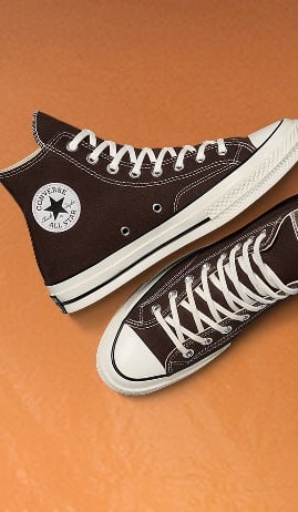 The image shows a pair of dark brown high-top Converse All Star sneakers with white laces and rubber soles on an orange background. One shoe displays the iconic circular Converse logo patch, while the other is shown from a top-down view.