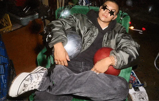 A person wearing a shiny bomber jacket and star-shaped sunglasses is reclining in a green armchair with legs crossed. They hold a silver and a red ball, and wear patterned high-top sneakers and multiple rings.