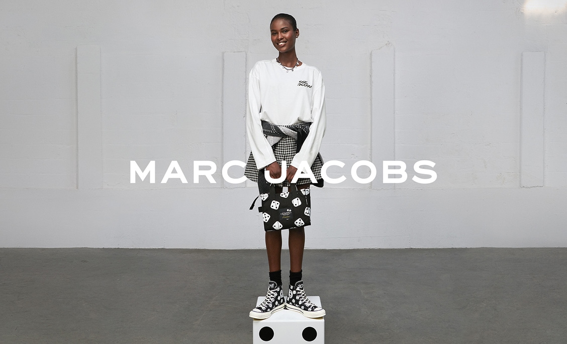A smiling model wearing a white long-sleeve top, patterned skirt, and black-and-white high-top sneakers stands on a white cube against a plain light-gray wall. Bold white text &ldquo;MARC JACOBS&rdquo; is overlaid across the center of the image.
